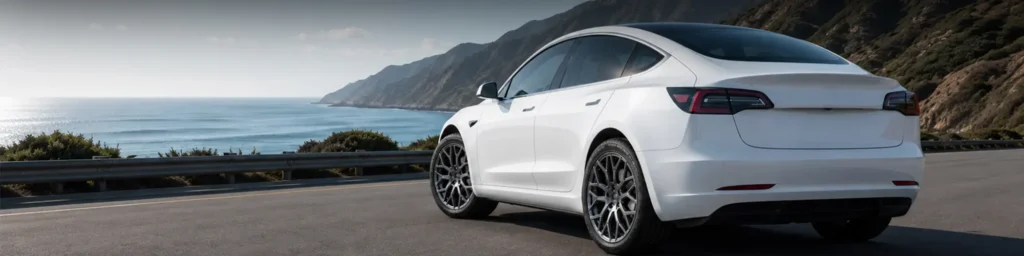 White electric vehicle driving along a coastal road with mountains and ocean